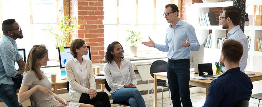 Man standing up talking with a group of people sitting and standing in an office listening.