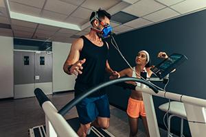 Man running on treadmill with fitness mask on while woman is looking on watching screen with results on it.